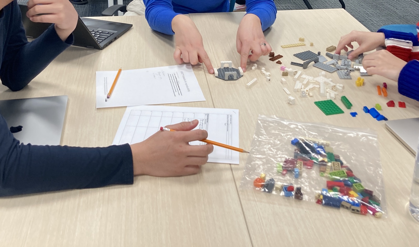 Students' hands building small LEGO models next to worksheets and a bag of bricks.