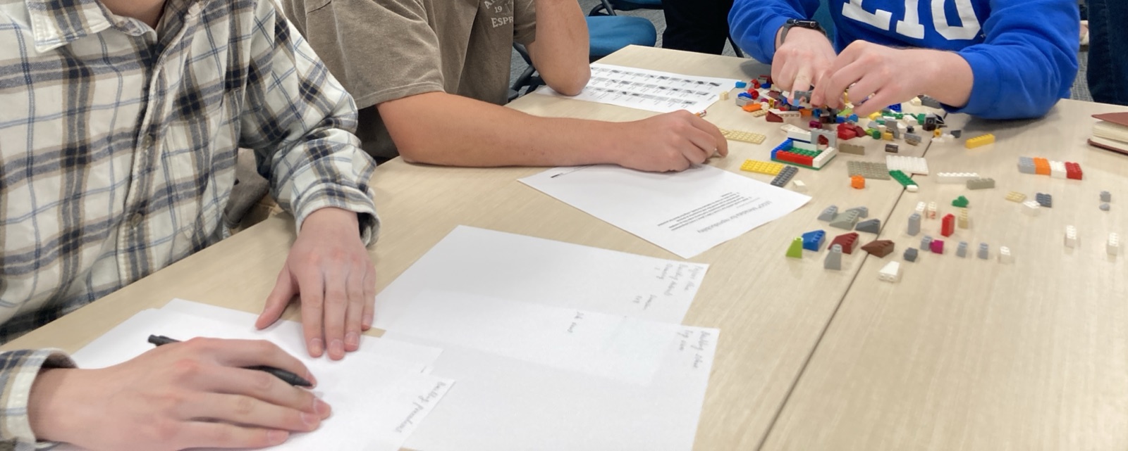 Students writing notes on worksheets beside their LEGO builds.